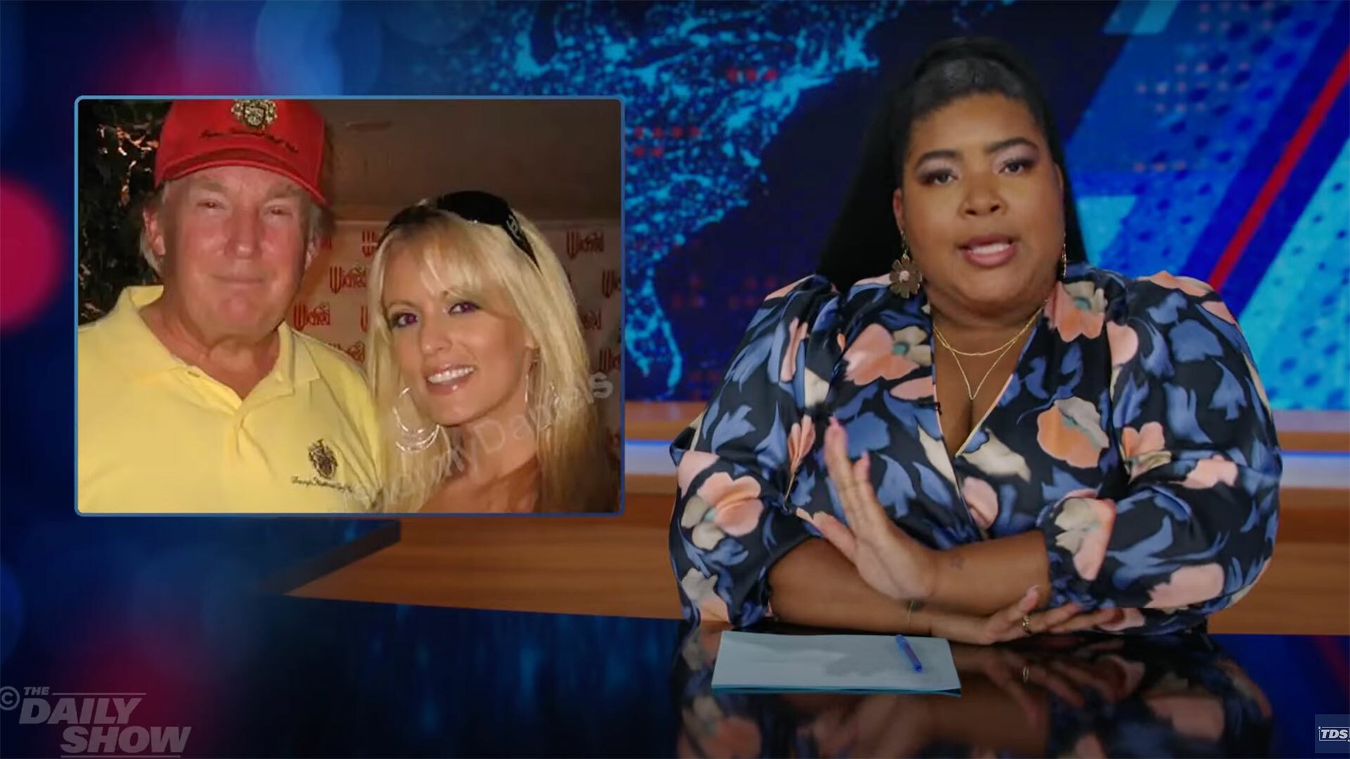 A woman sits at a talk show desk. In the top-left is an image of a man and a woman.