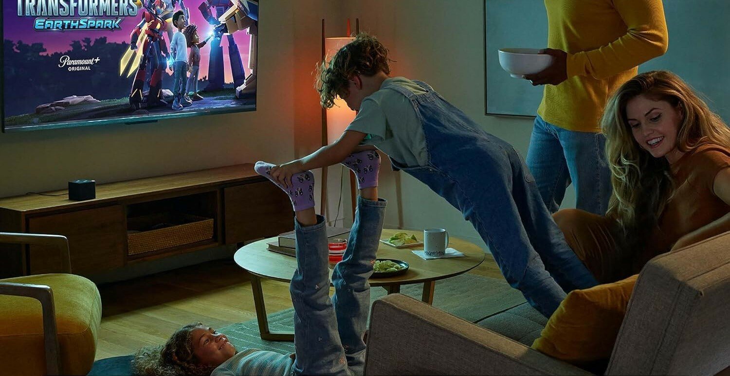 a family watches a film on their living room tv with an amazon fire tv cube device sitting on the entertainment system under the TV