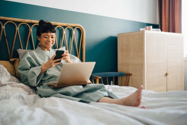 Woman using phone and laptop on bed.