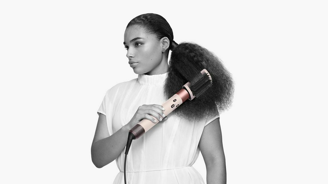 a person stands against a white background while using the dyson airwrap complete long hair styling tool with a brush attachment