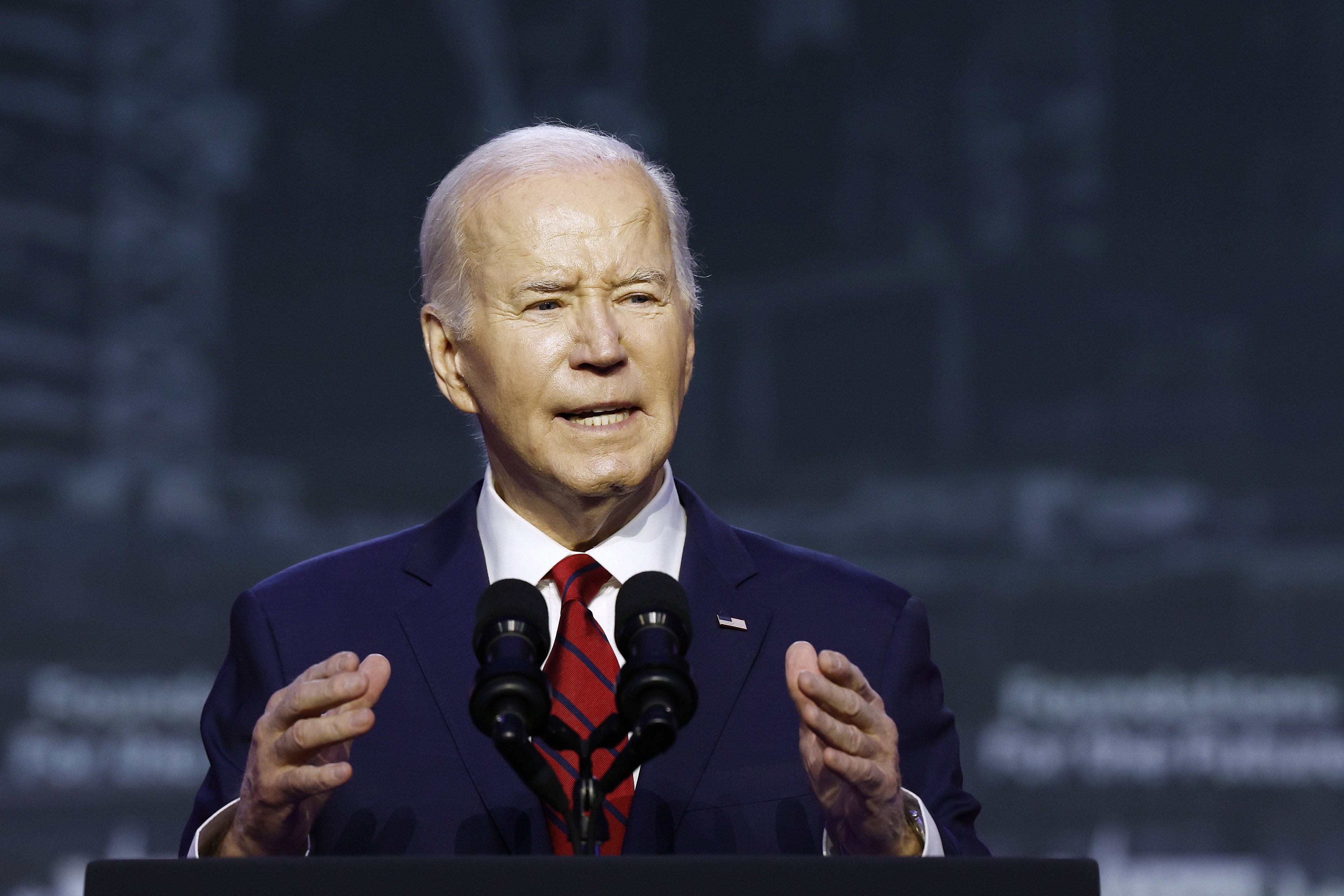 President Joe Biden standing behind a lectern in a navy suit and red tie. 