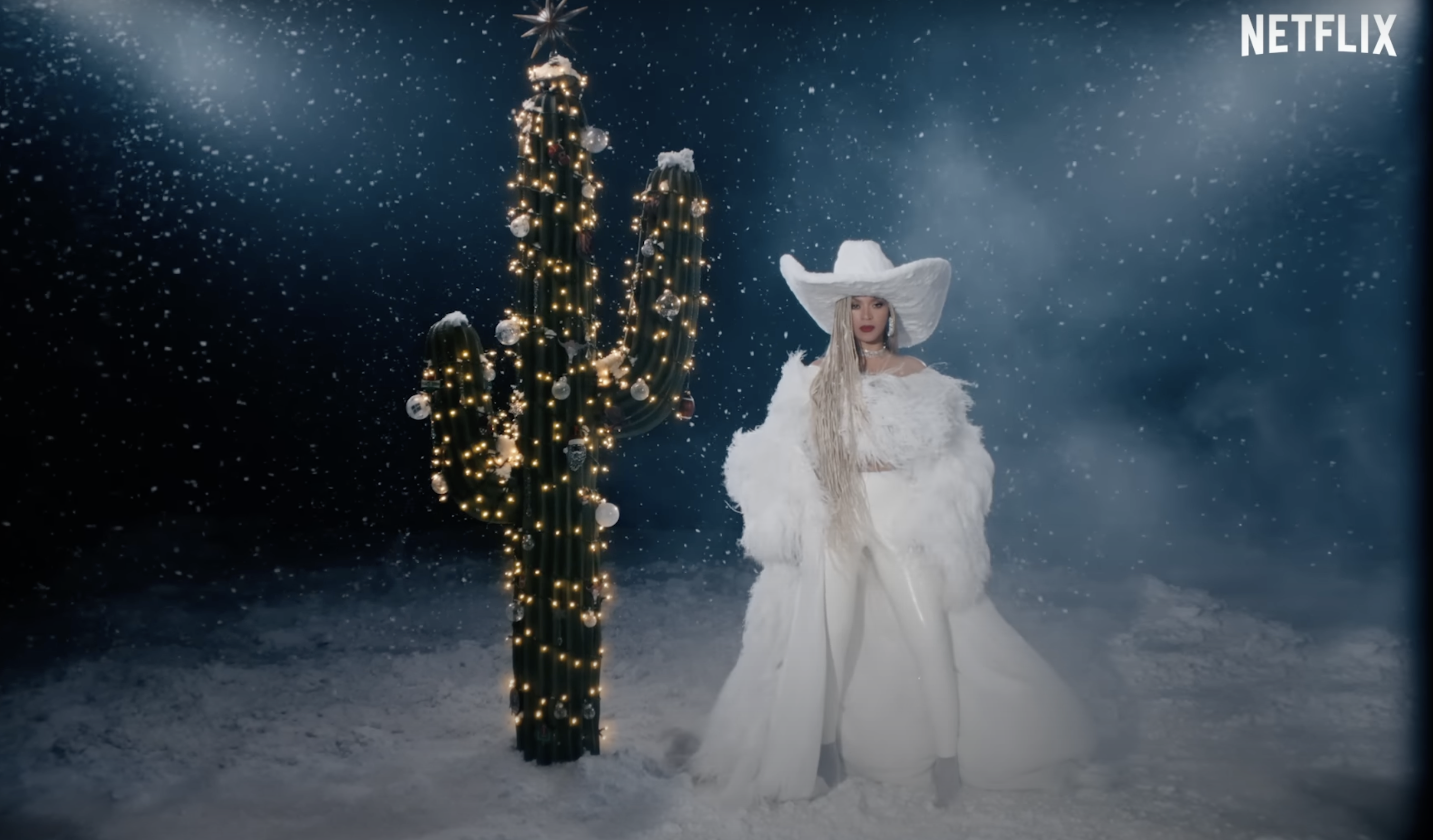 Beyonce dressed in all white standing next to a cactus Christmas tree in the snow.